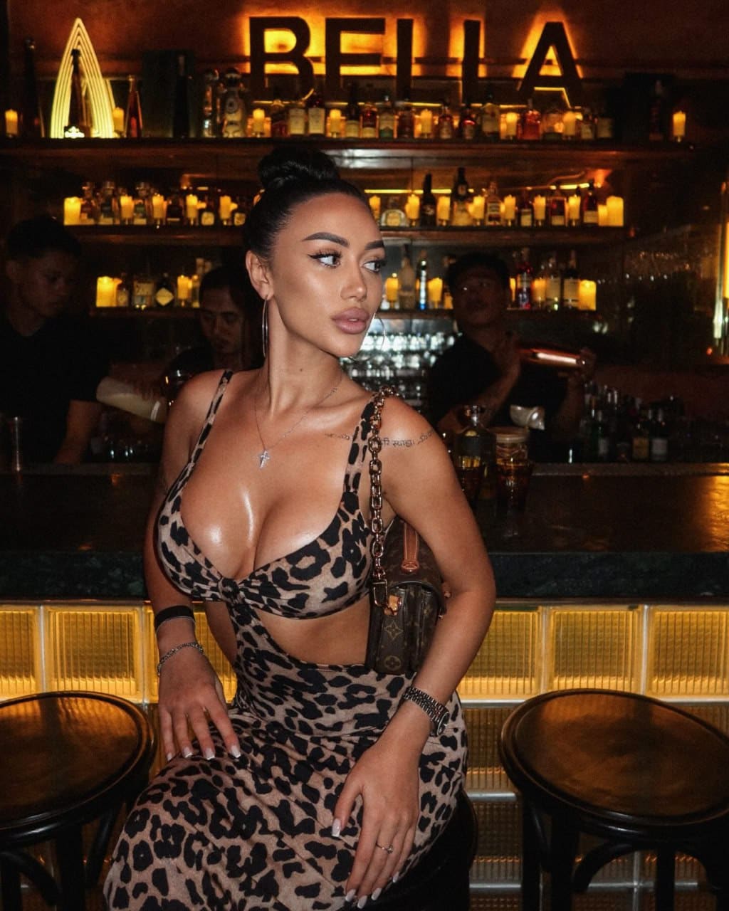 A woman sits at a bar counter in a leopard-print cut-out dress, with illuminated bottle shelves behind her.