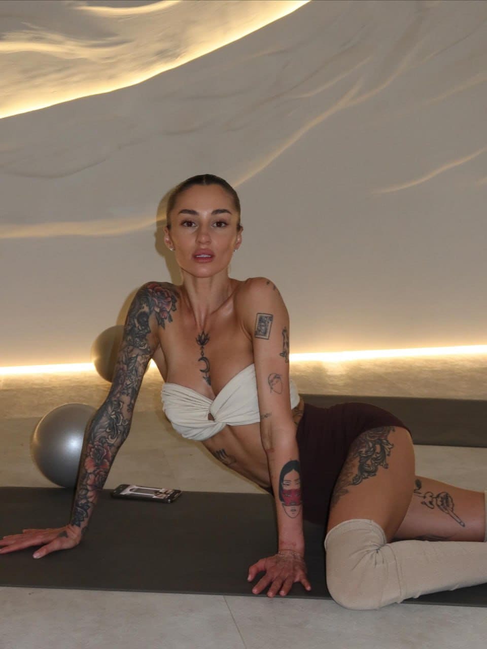 A tattooed woman sits on a black mat in a white strapless top and dark shorts, with an exercise ball beside her in a curved room.