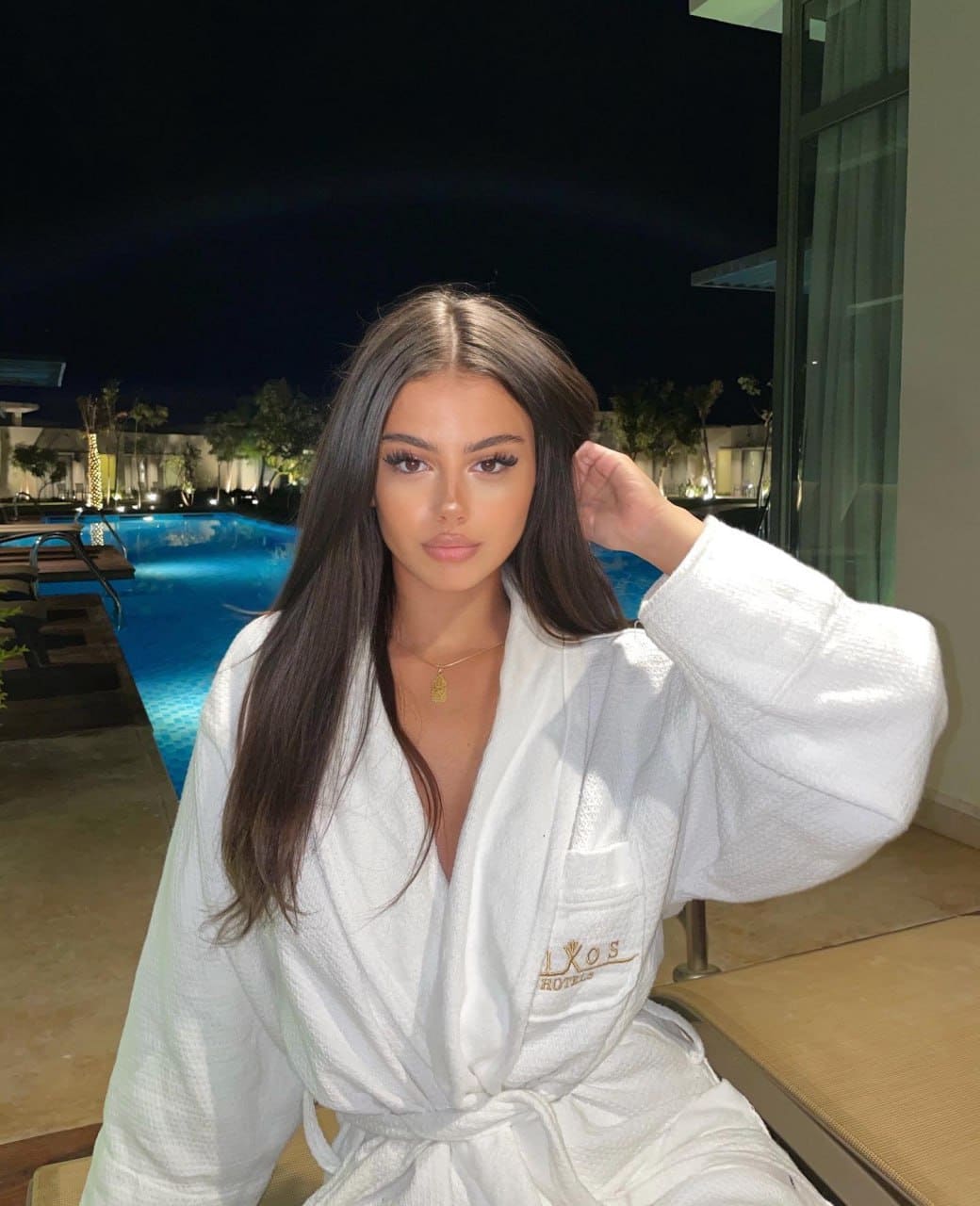 A woman sits by a lit swimming pool at night, wearing a white waffle-knit bathrobe tied at the waist, with one hand raised to her hair.