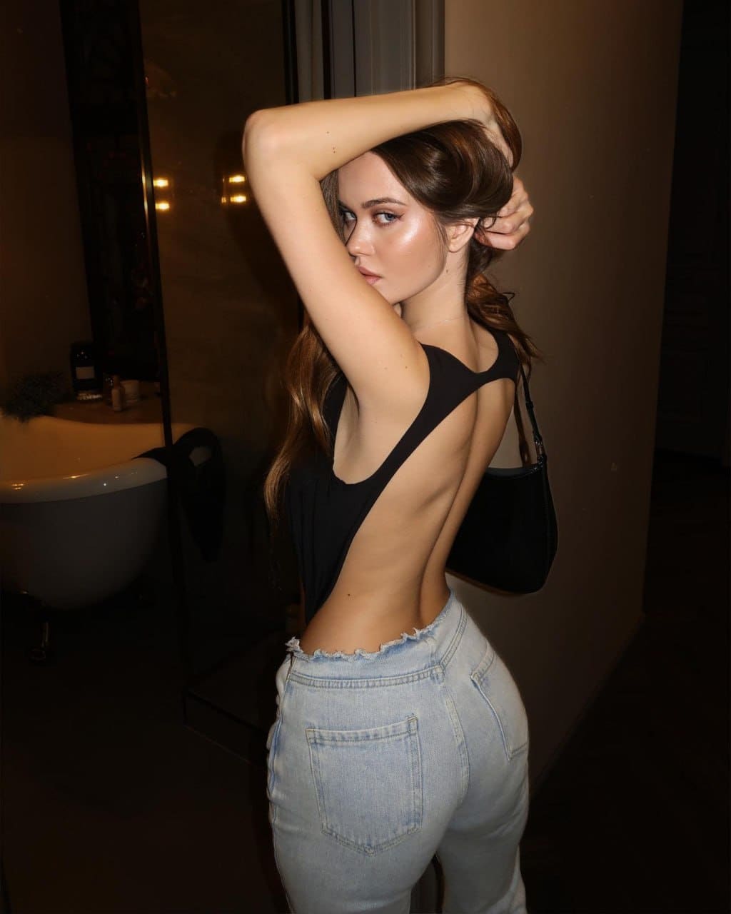 In profile, a woman holds her hair back, wearing a backless black bodysuit, jeans, and a shoulder bag in a dim interior.