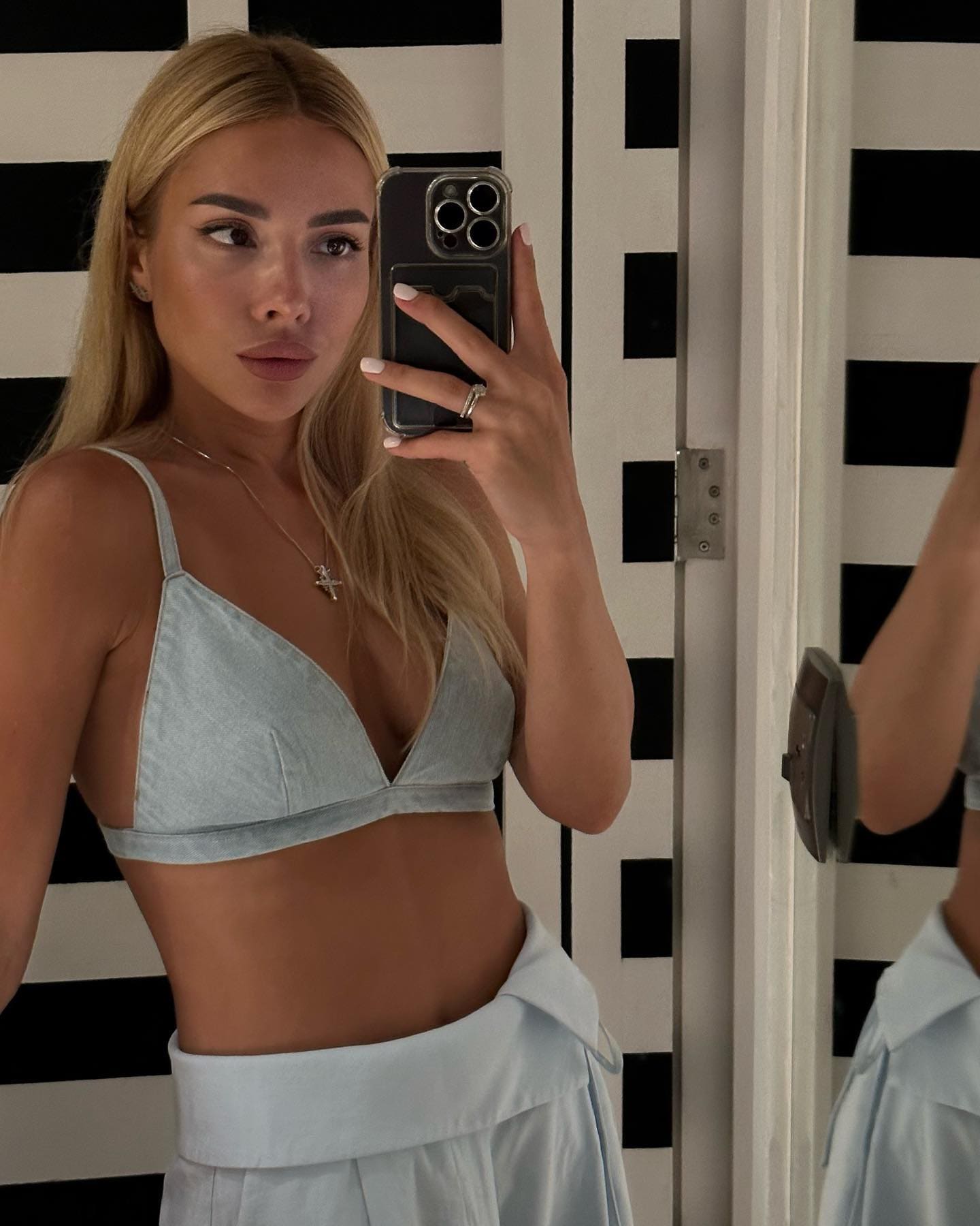 A blonde woman in a light blue bralette and high-waisted skirt takes a mirror selfie against a checkered wall.