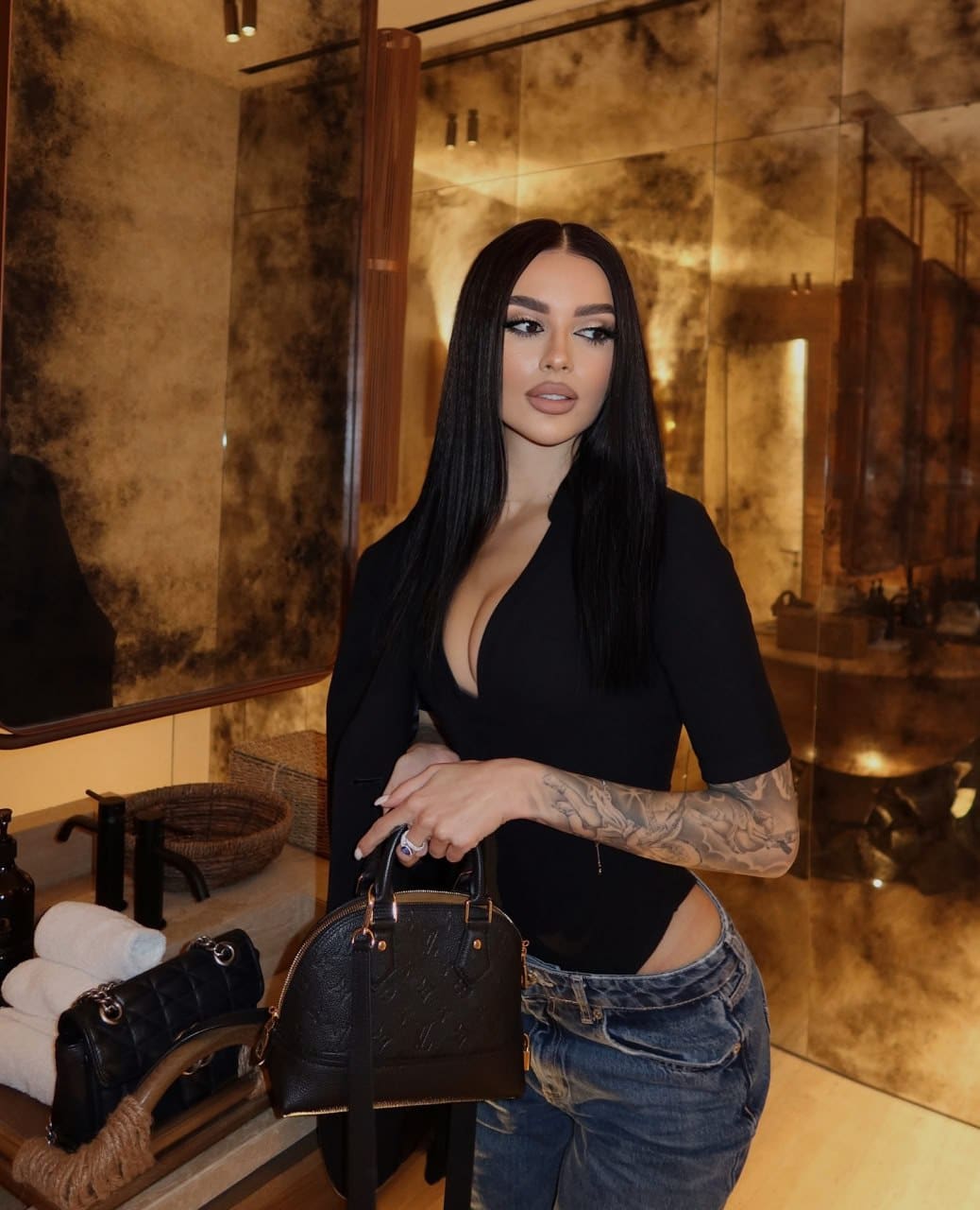 Vasilisa stands in a bathroom holding a dark handbag, wearing a black V-neck top and blue jeans, with a tattooed left arm.