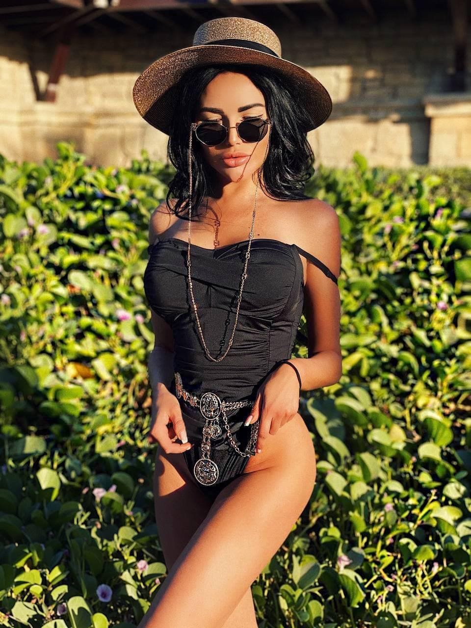 A woman stands before green foliage wearing a straw hat, round sunglasses, and a black strapless bodysuit with a decorative belt.