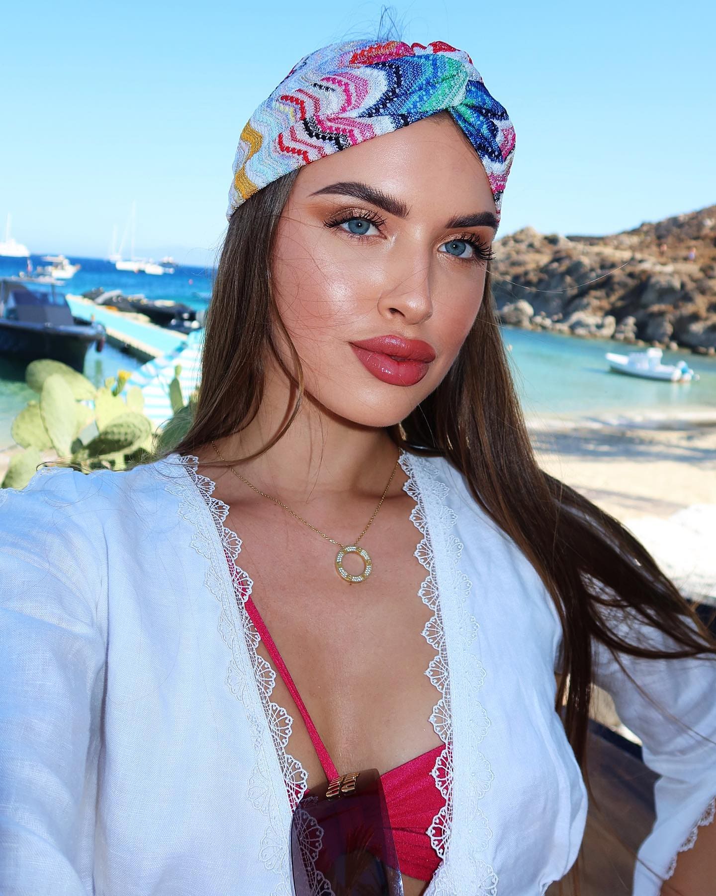 Woman in a white lace cover-up and patterned headscarf poses over a red bikini with a harbor background.