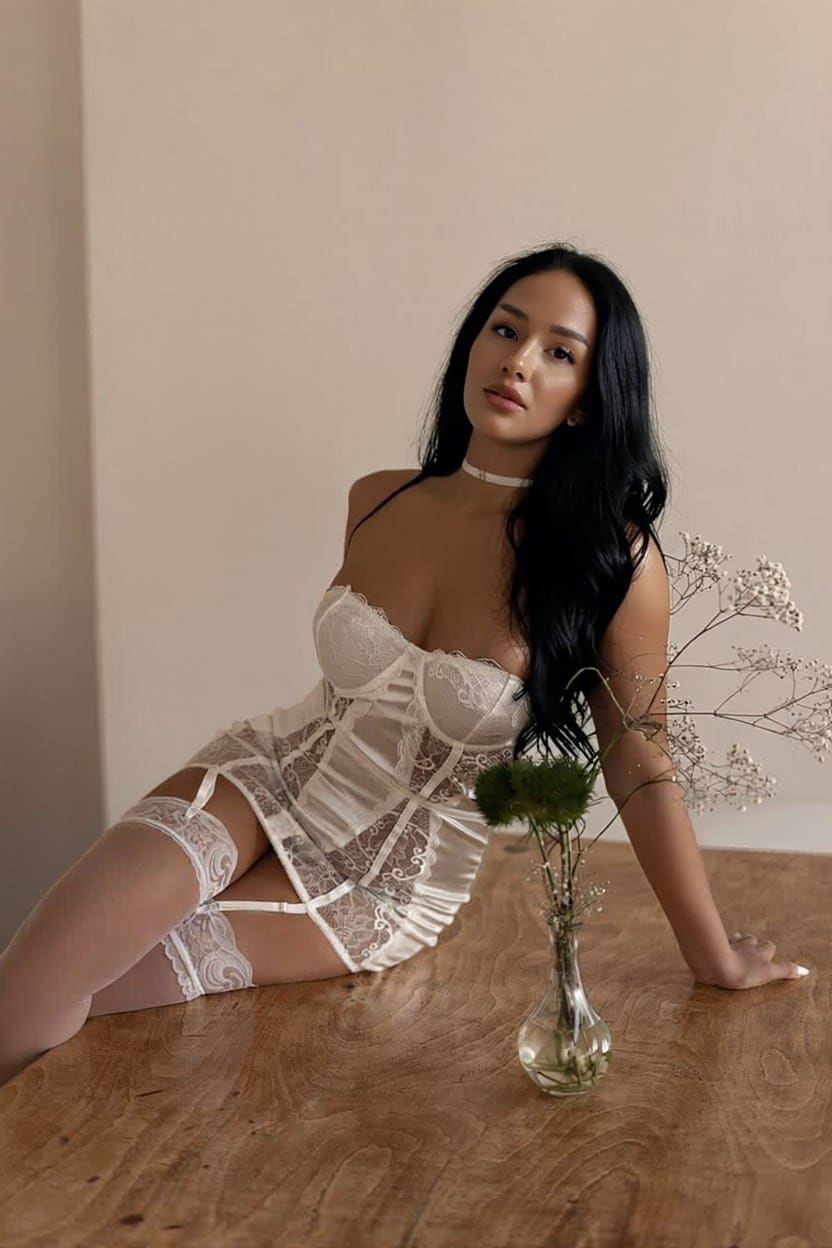 Reclining on a wooden table, she wears a white lace corset dress with garter straps and sheer stockings next to dried flowers.