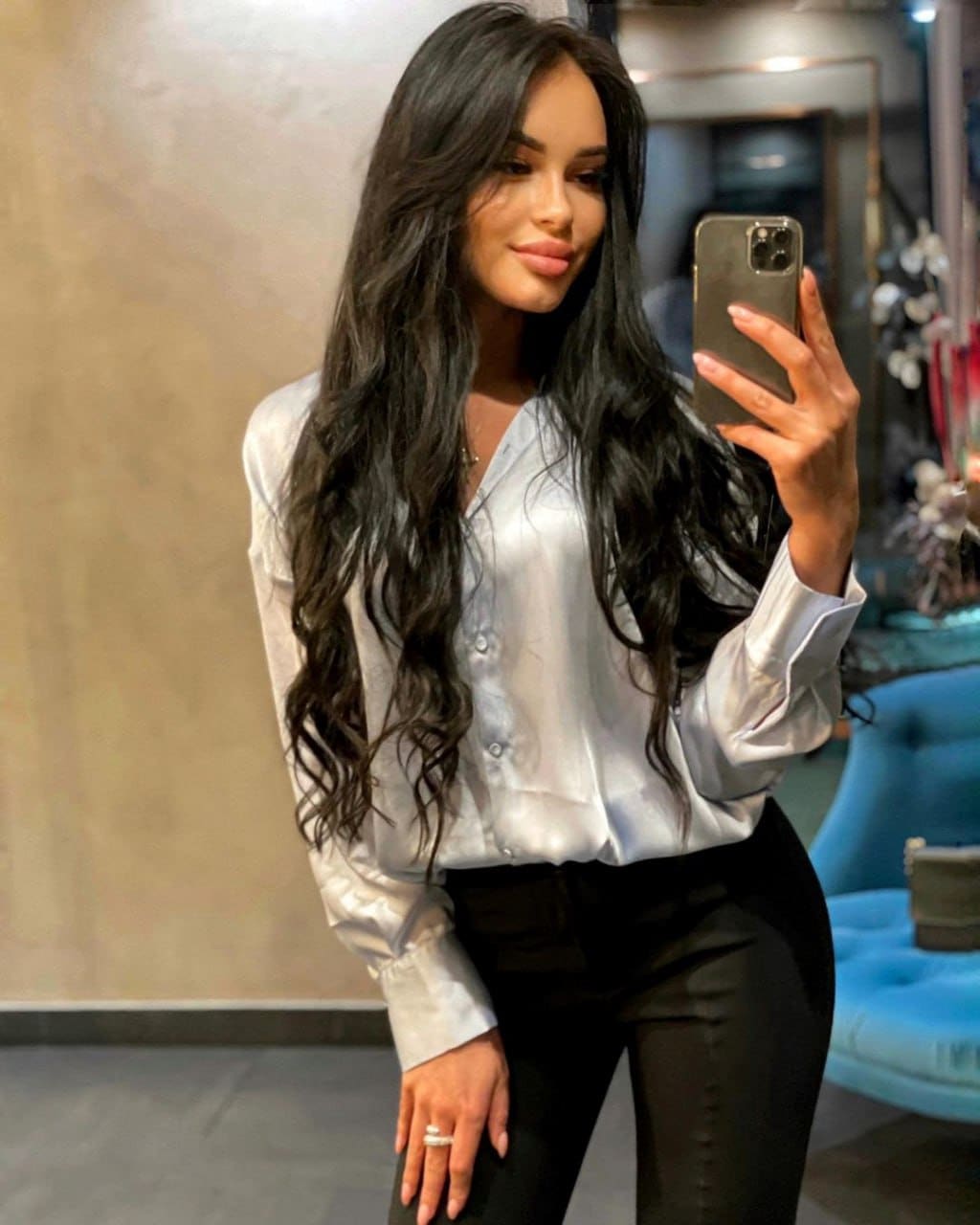 Mirror selfie of a woman in a white satin shirt and black trousers, with a blue chair visible in the background of the room.