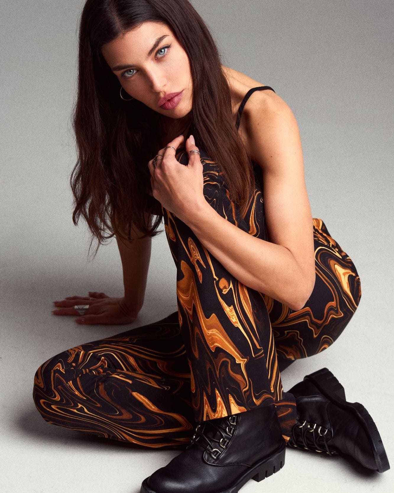 A woman kneels on a gray floor in a black and orange jumpsuit and lace-up boots, one hand on the ground.
