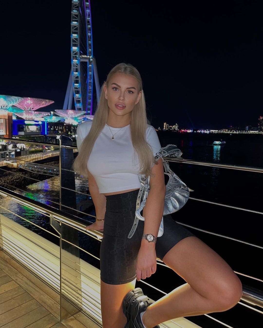A blonde woman stands on a night promenade in a white cropped top, black mini skirt, and sneakers, with a silver bag and lit Ferris wheel behind.