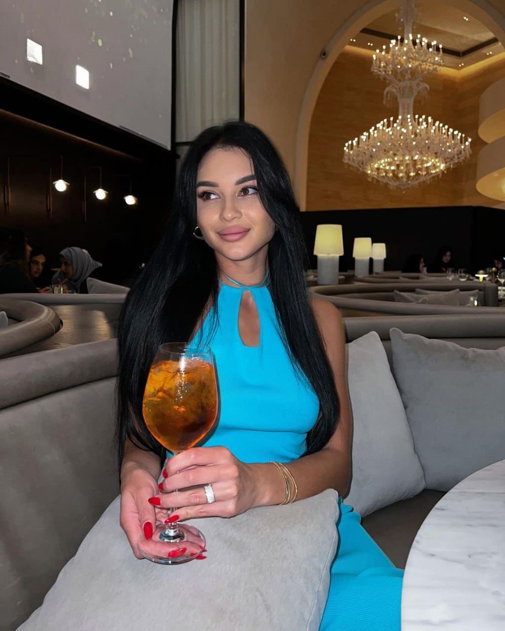 Lara sits on a grey sofa in a restaurant, wearing a turquoise keyhole dress and holding a glass of amber drink with ice.