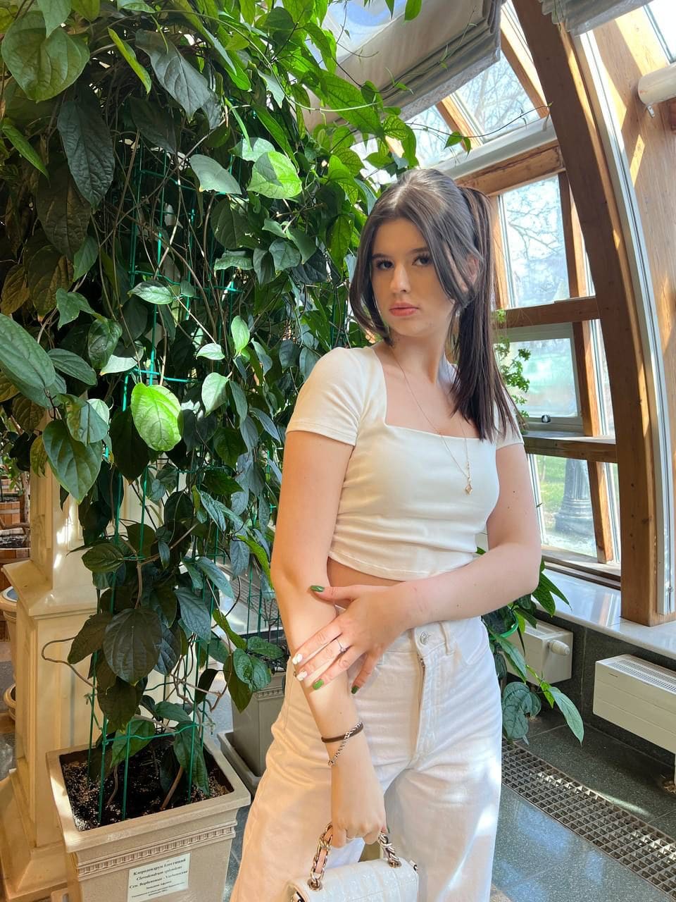A woman with a high ponytail stands indoors in a white crop top and pants, holding a small handbag next to a large potted plant in a sunlit room.