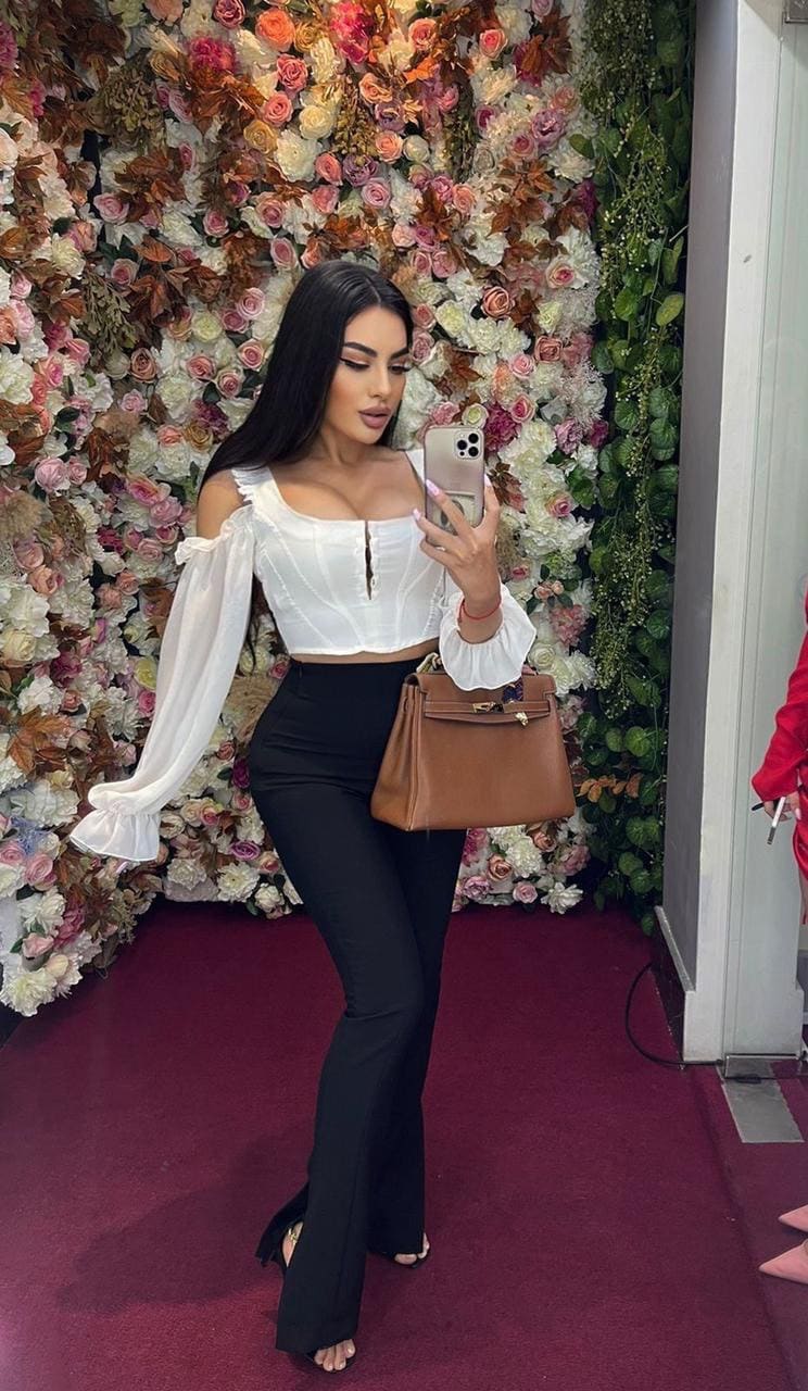 A woman in a white off-shoulder top and black flared trousers takes a mirror selfie against a floral wall on a red carpet.