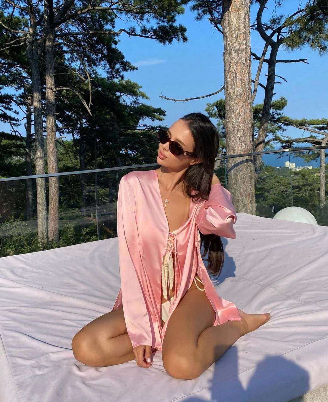 A woman wearing sunglasses sits cross-legged on a white sheet outdoors, dressed in an open pink satin robe over a light-colored bikini, with trees behind.