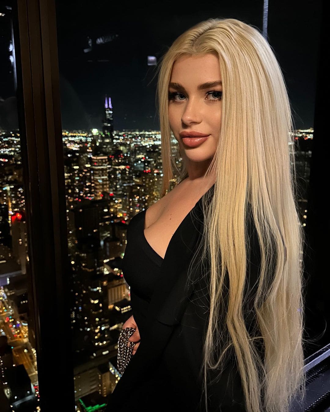 A woman in a black V-neck dress stands by a window at night, holding a beaded clutch, with a lit city skyline in the background.