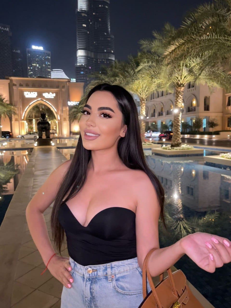A woman in a black strapless top and light-wash jeans stands outdoors at night near a lit cityscape and reflecting pool.