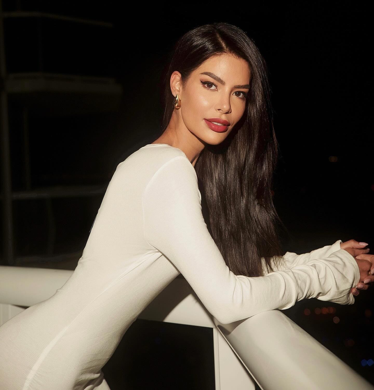 A woman in a fitted white long-sleeve top and matching pants leans on a white railing at night, set against a dark background.
