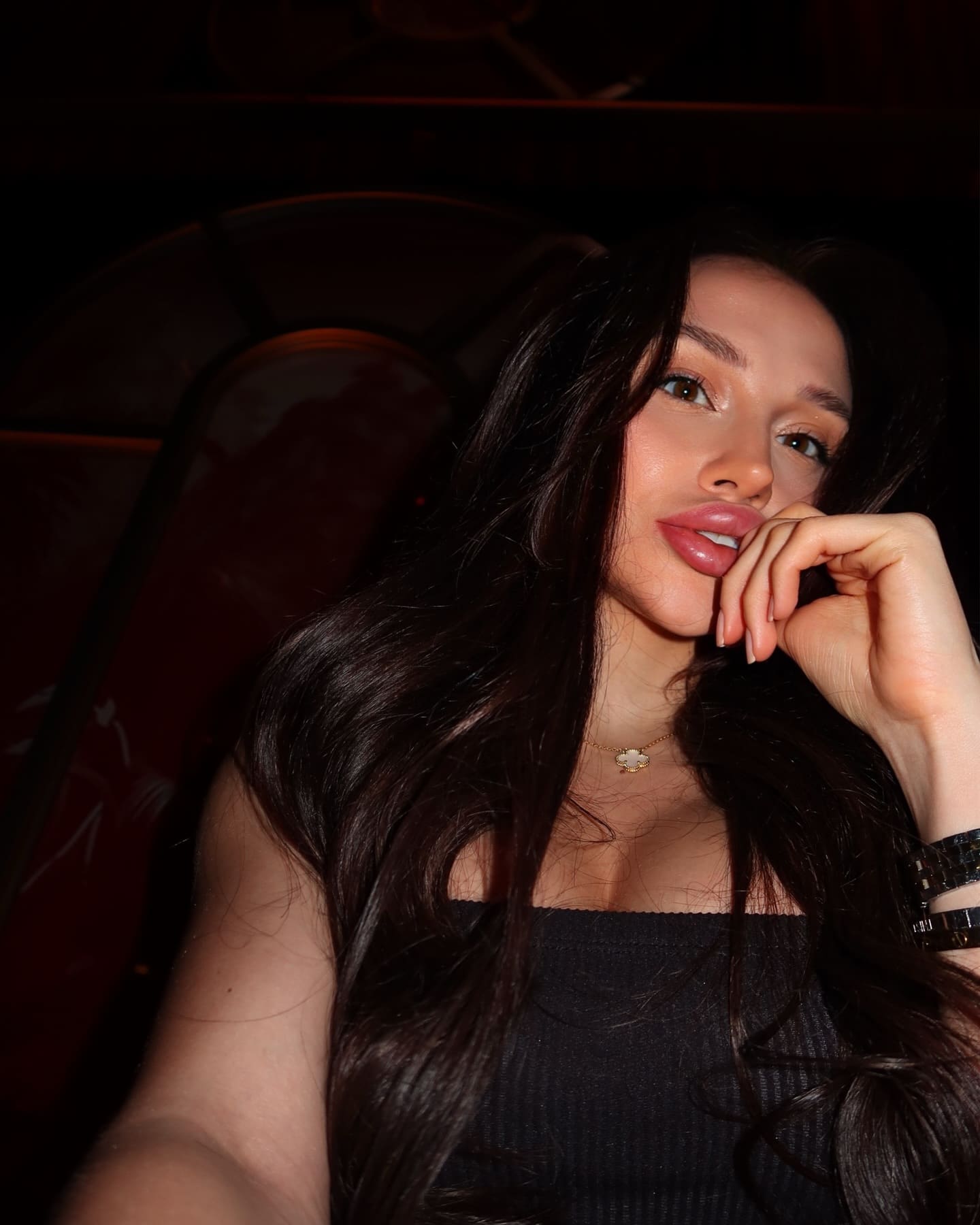 A woman in a black off-the-shoulder top rests her chin on her hand, seated in a dim room with a dark red chair behind her.