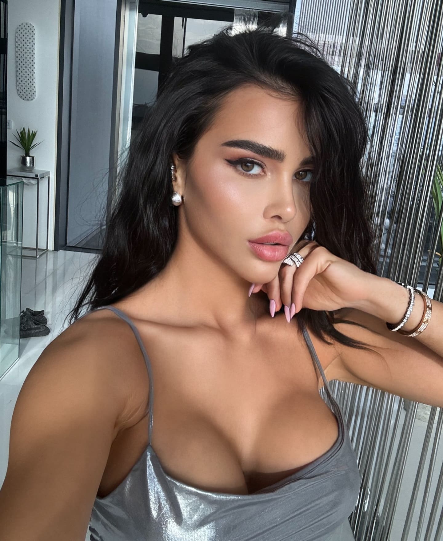A close-up selfie shows a woman with long dark hair in a silver metallic camisole, hand near her chin, with glass panels and a plant in the background.