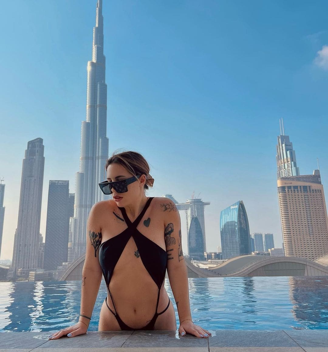 A tattooed woman in a black strappy swimsuit and sunglasses leans on a pool edge, with a city skyline of skyscrapers visible under a clear blue sky.