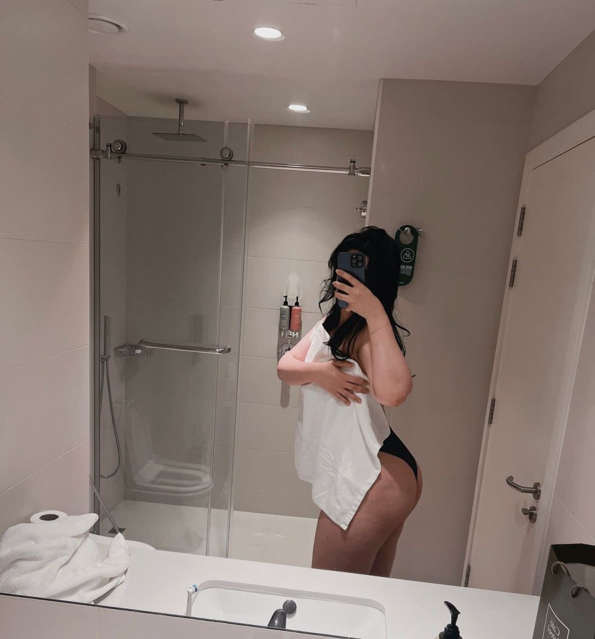 Bathroom mirror selfie wearing a loose white t-shirt and black underwear, with a glass shower enclosure behind.