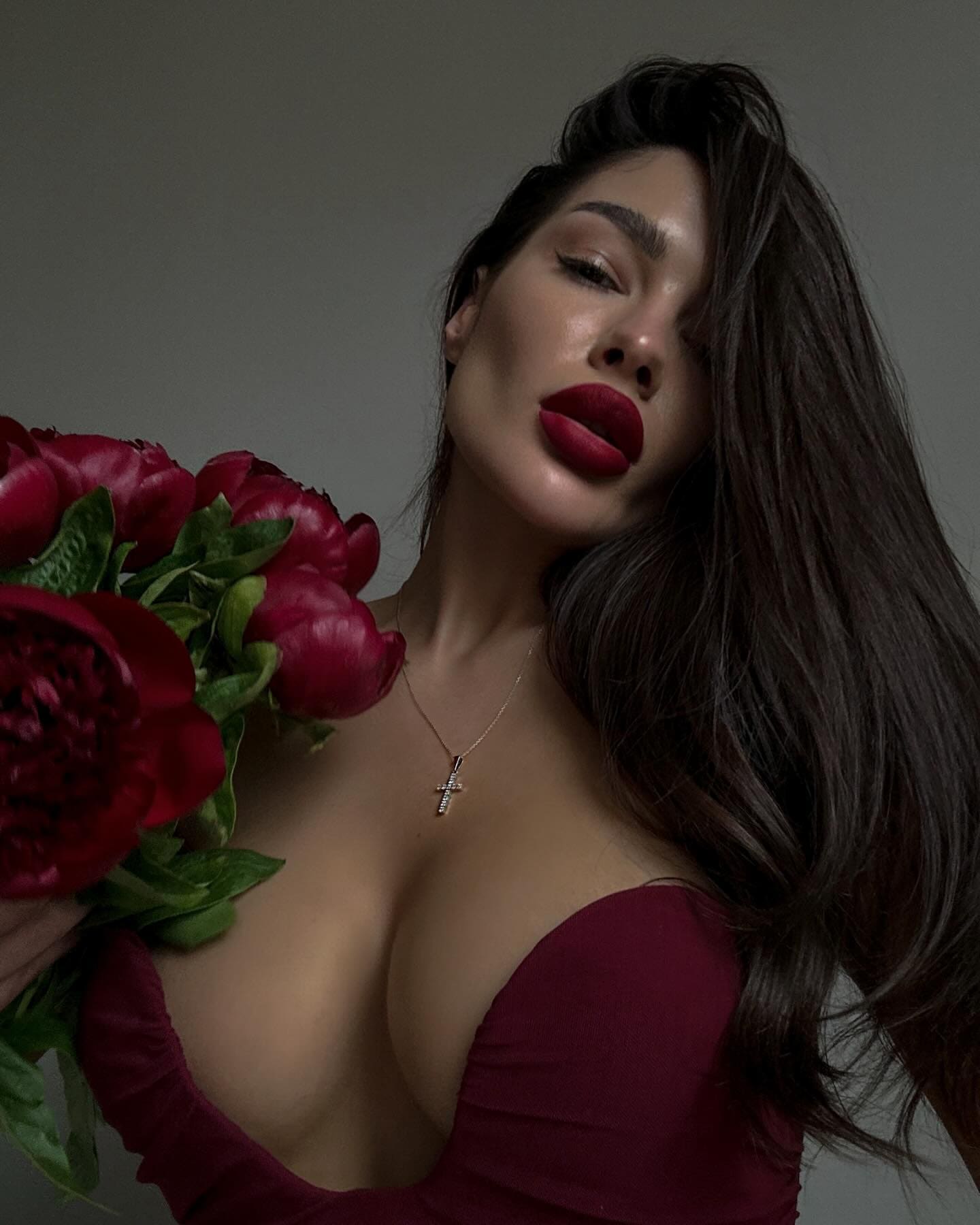 A woman with red lipstick holds red peonies while wearing a deep-plunging maroon top against a plain grey background.