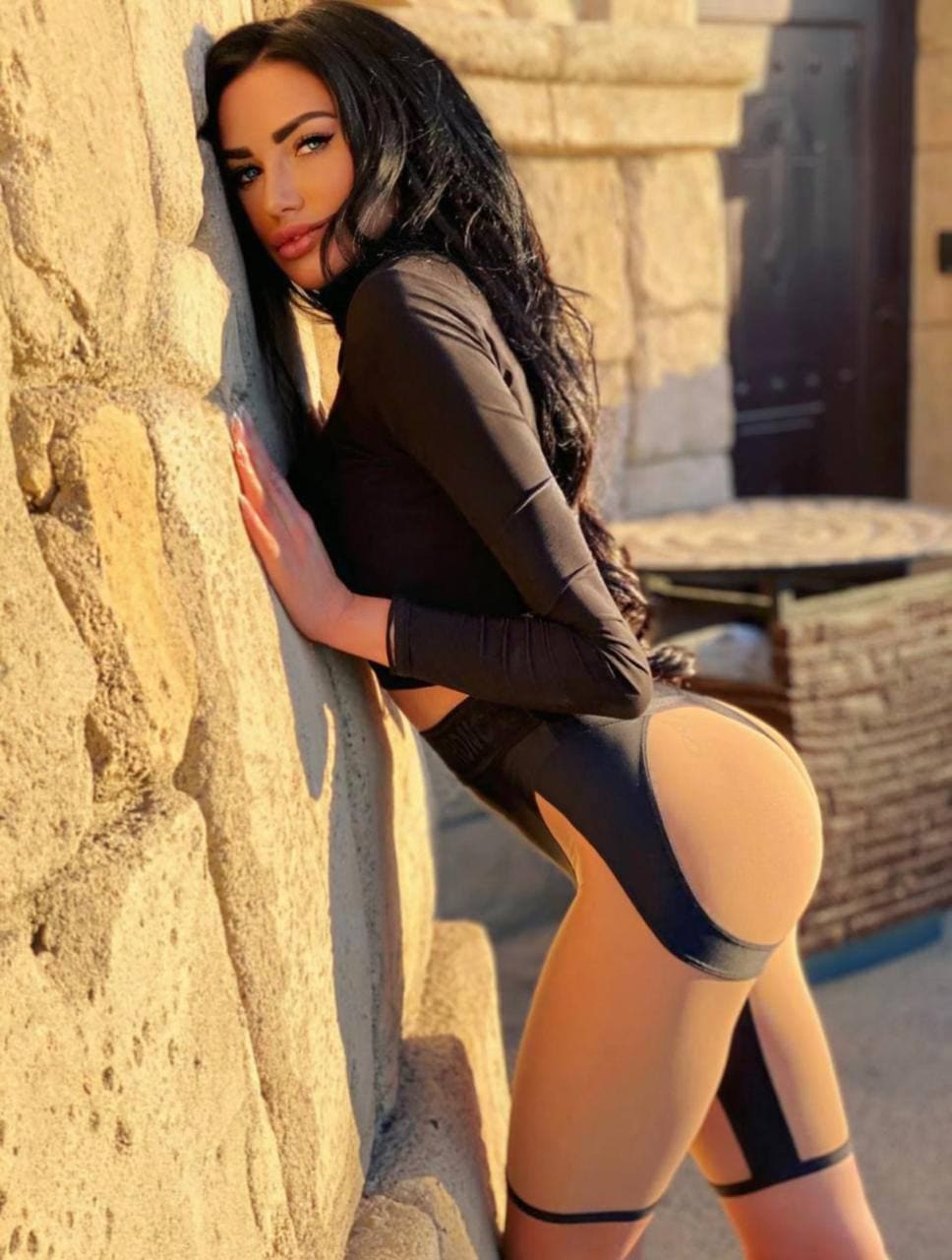 A woman leans against a rough stone wall outdoors, wearing a black long-sleeved crop top and a black bodysuit with thigh straps.