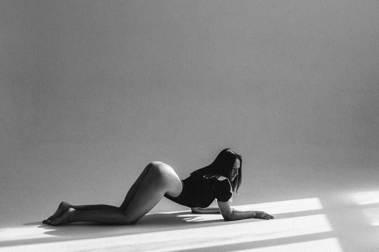A woman in a short dark dress lies prone on a light studio floor, arms extended forward, head turned.