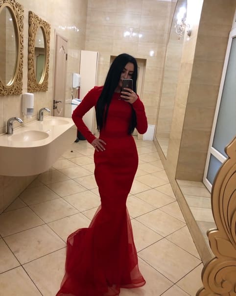 Mirror selfie in a tiled bathroom showing a long red gown with a fitted bodice, phone held to face and hand on hip.
