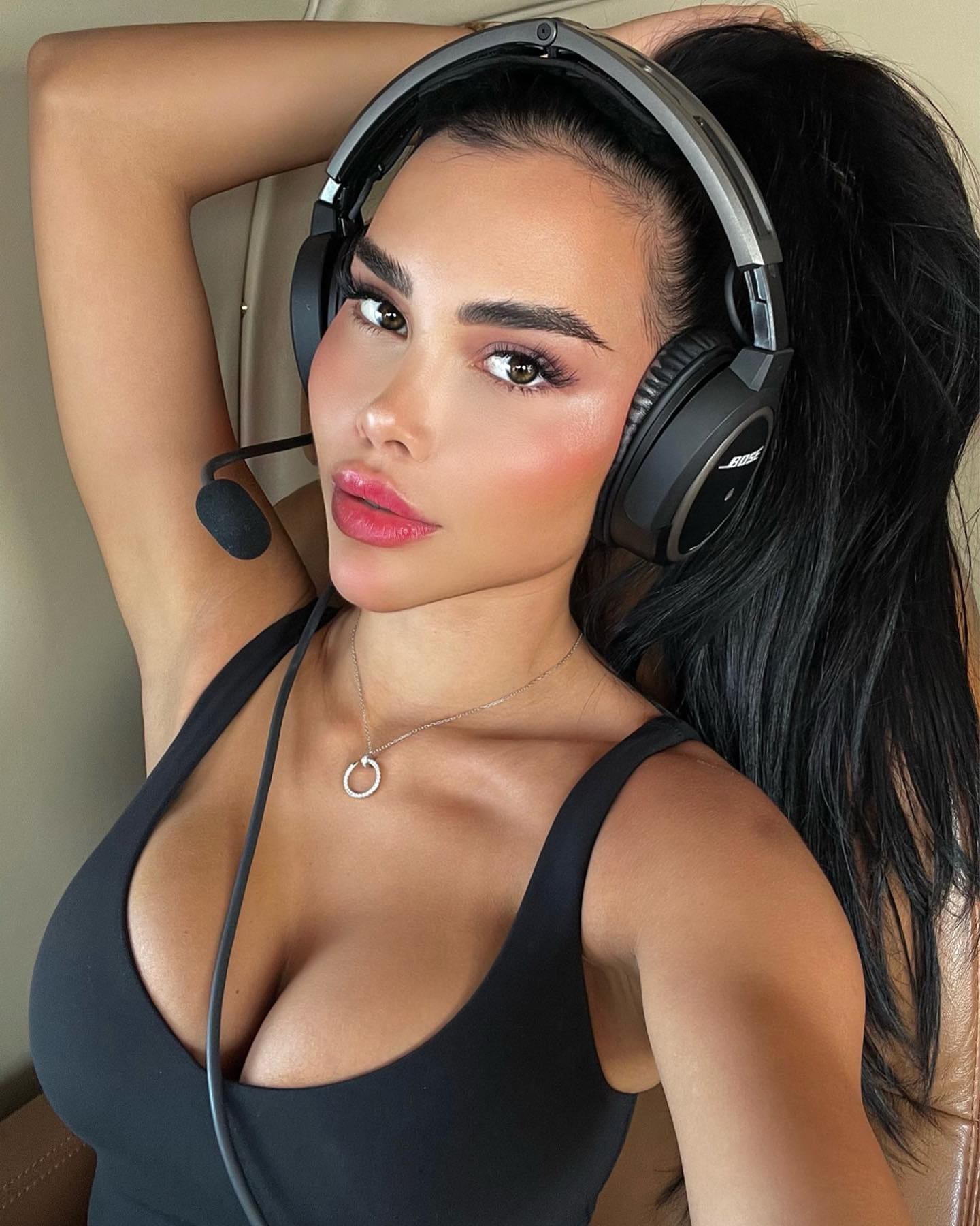 A woman with a high ponytail wears a black low-cut tank top and holds over-ear headphones to her head while seated against a tan leather surface.