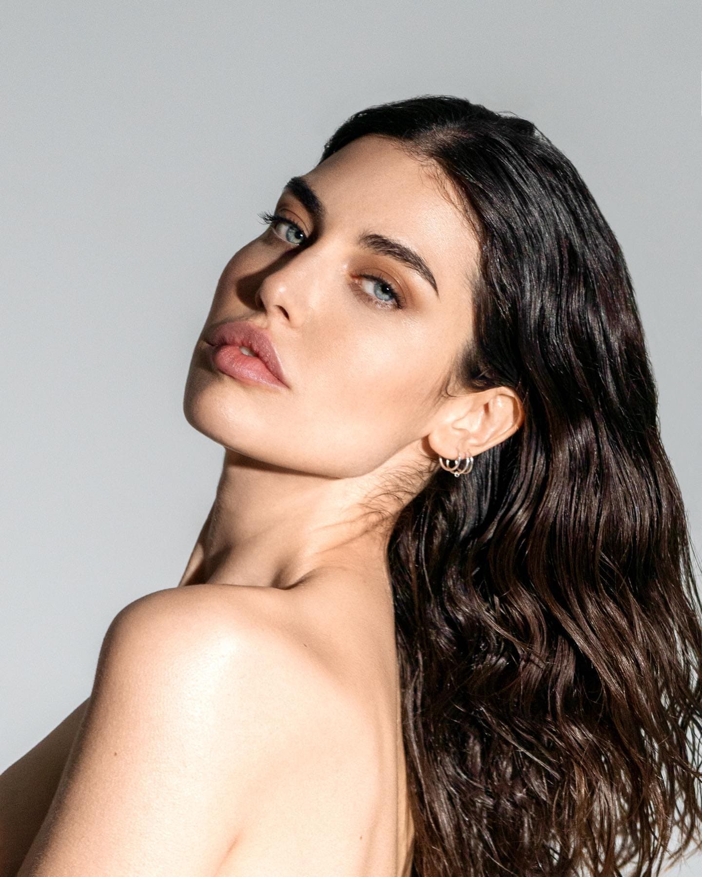 A bare-shouldered woman with hoop earrings tilts her head to look over her shoulder at the camera.