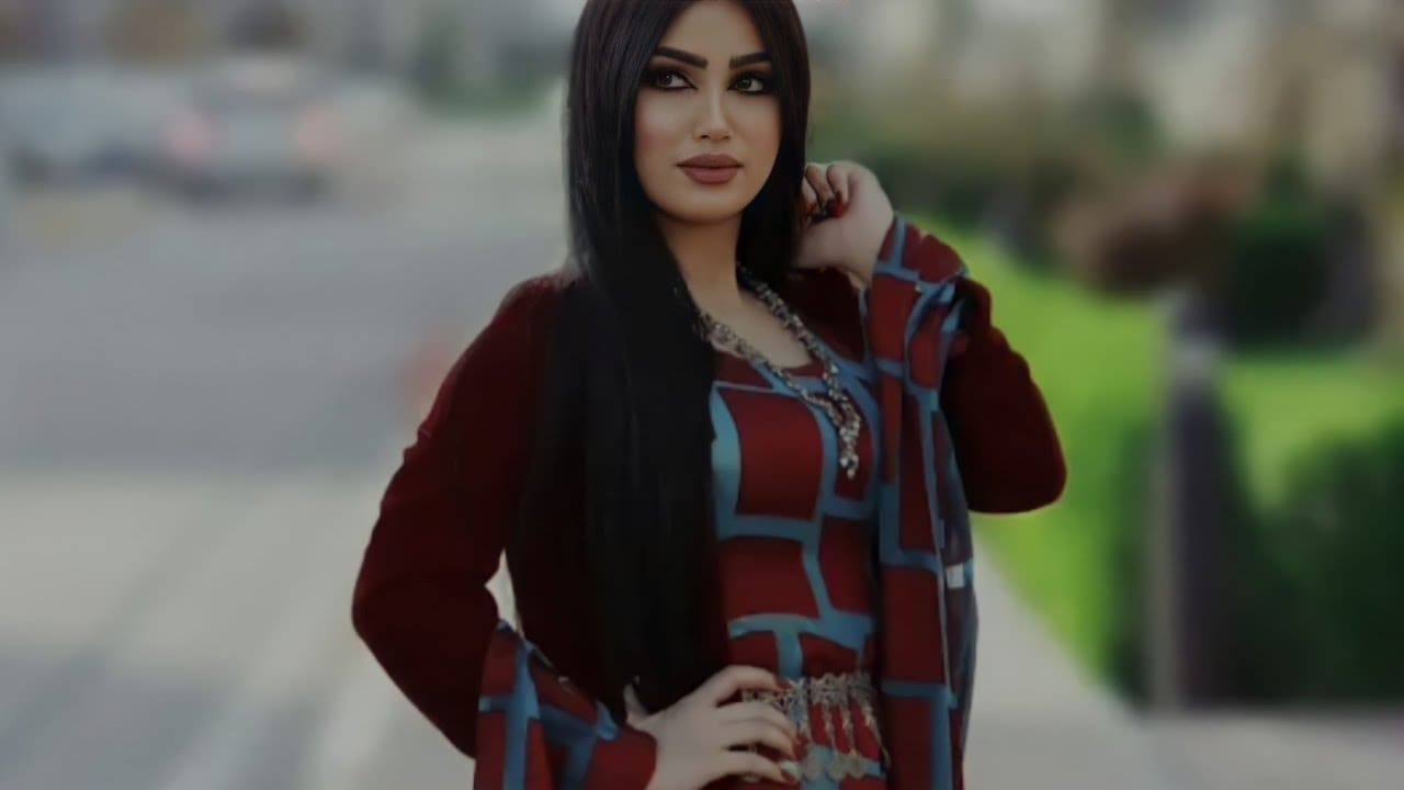 Outdoors in a blurred street, wearing a maroon and blue patterned top with cardigan, one hand raised to her hair.