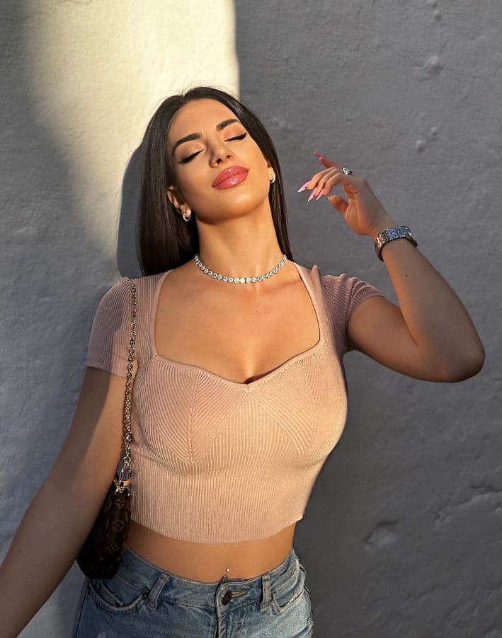 Polina stands against a textured wall in warm light, wearing a beige crop top, jeans, and jewelry, with one hand raised near her face.
