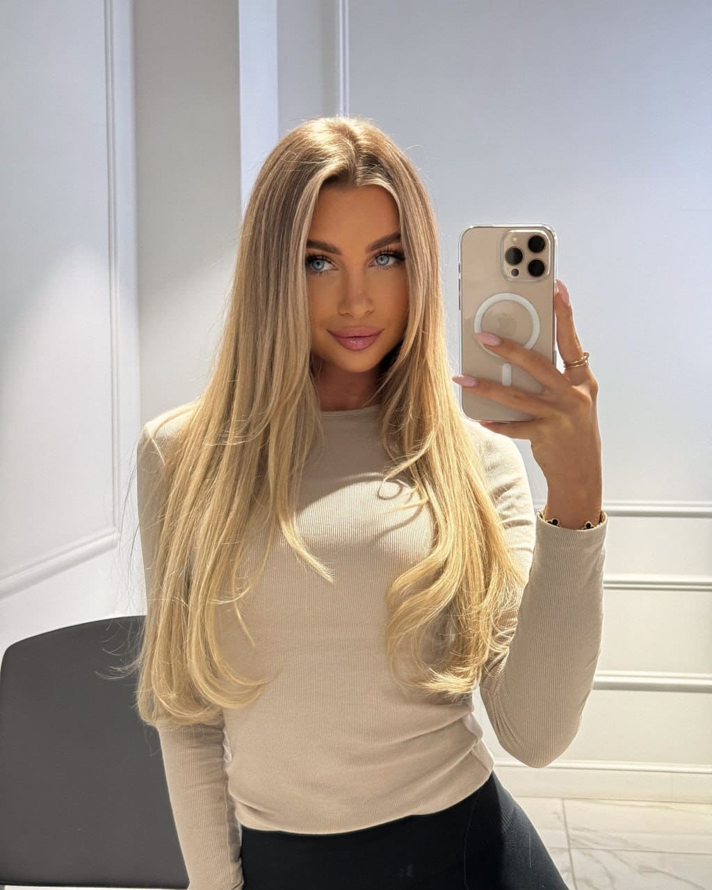A mirror selfie shows a woman in a fitted beige long-sleeved top and dark bottoms against white paneled walls.