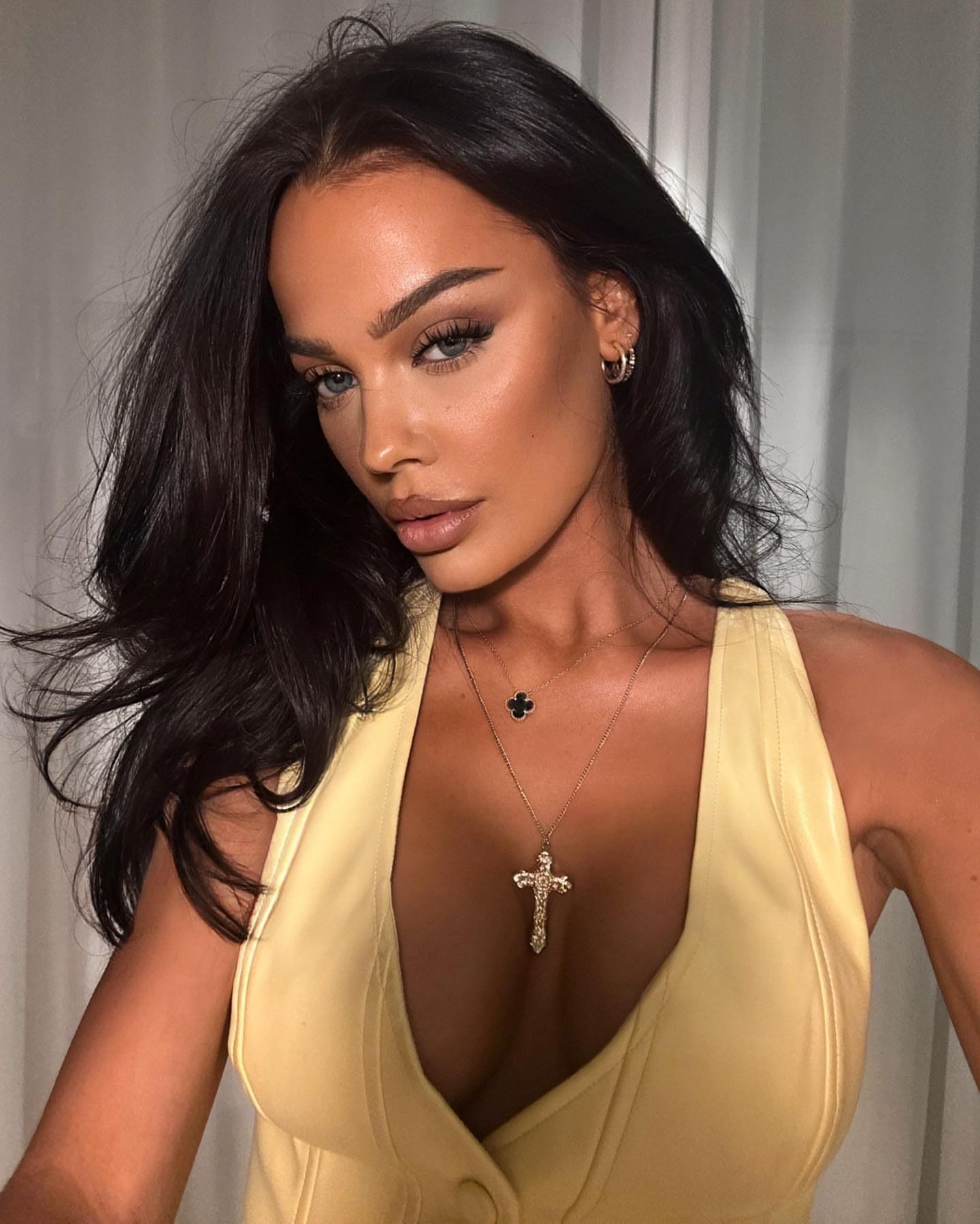 Malma with long dark wavy hair in a pale yellow sleeveless V-neck top and gold necklaces, posing against a light gray curtain.