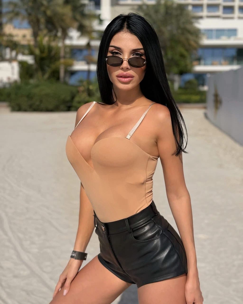 A woman in sunglasses, a beige strapless bodysuit, and black leather shorts poses outdoors with a hand on her thigh, buildings and greenery behind her.