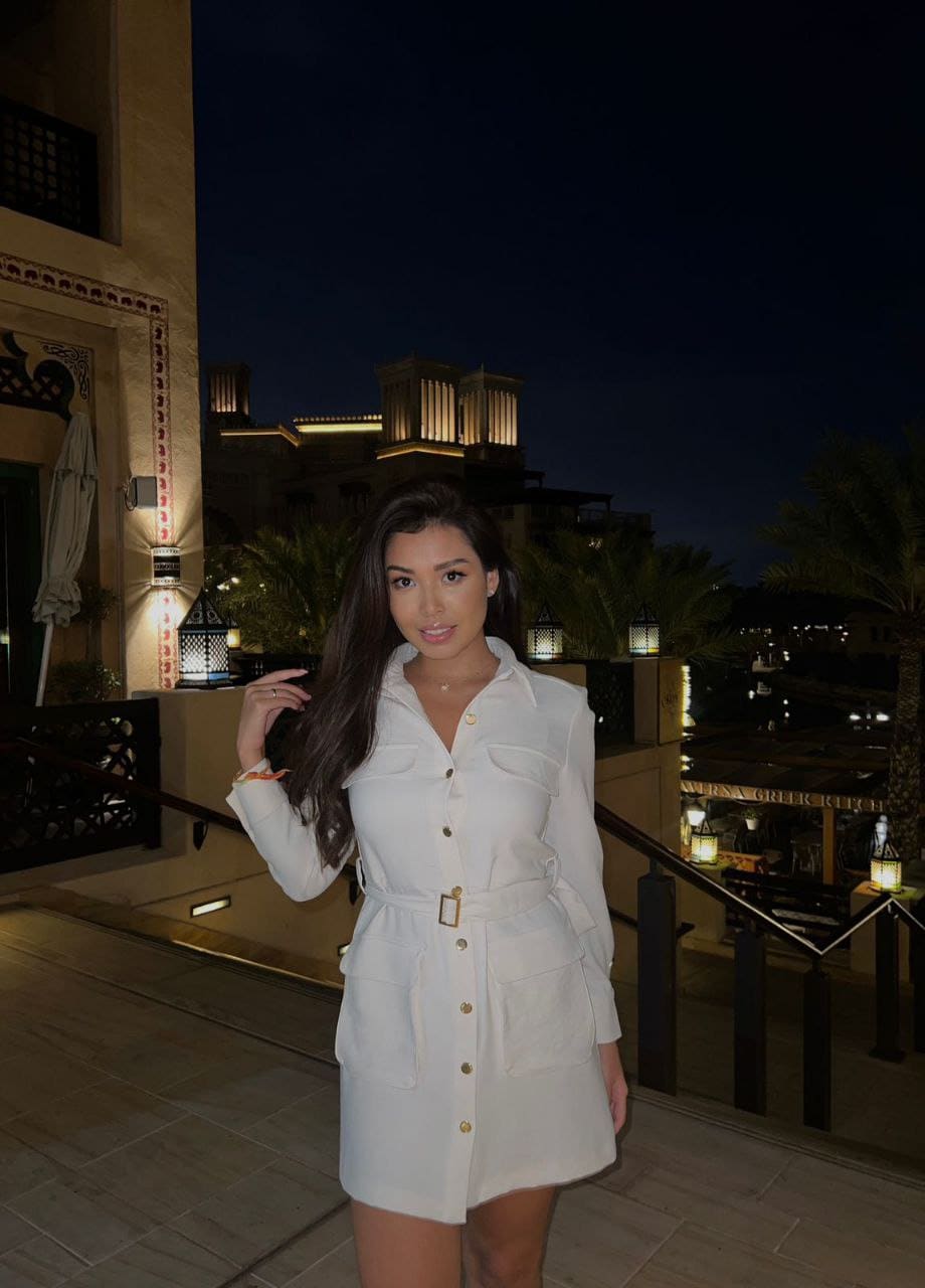 A woman in a white belted shirt dress stands on a night terrace, one hand near her hair, lit by ambient lanterns and city lights.