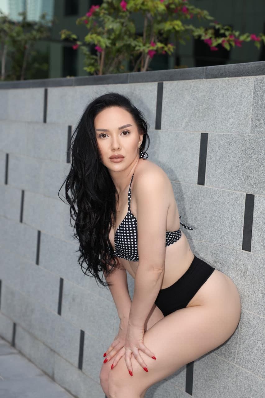 Seated outdoors against a gray tiled wall, she wears a black and white polka-dot halter swimsuit top and black high-waisted bottoms.