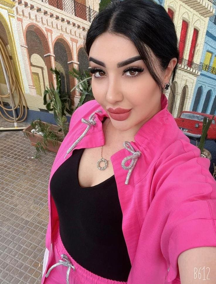 Medina stands before a colorful facade in a pink zip-up jacket and black top, wearing a pendant necklace and earrings.