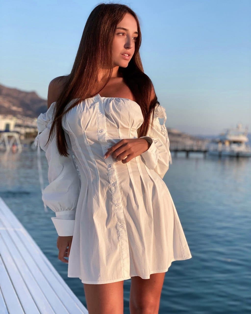A woman in a white off-the-shoulder dress stands on a deck by water, hand on chest, looking aside.