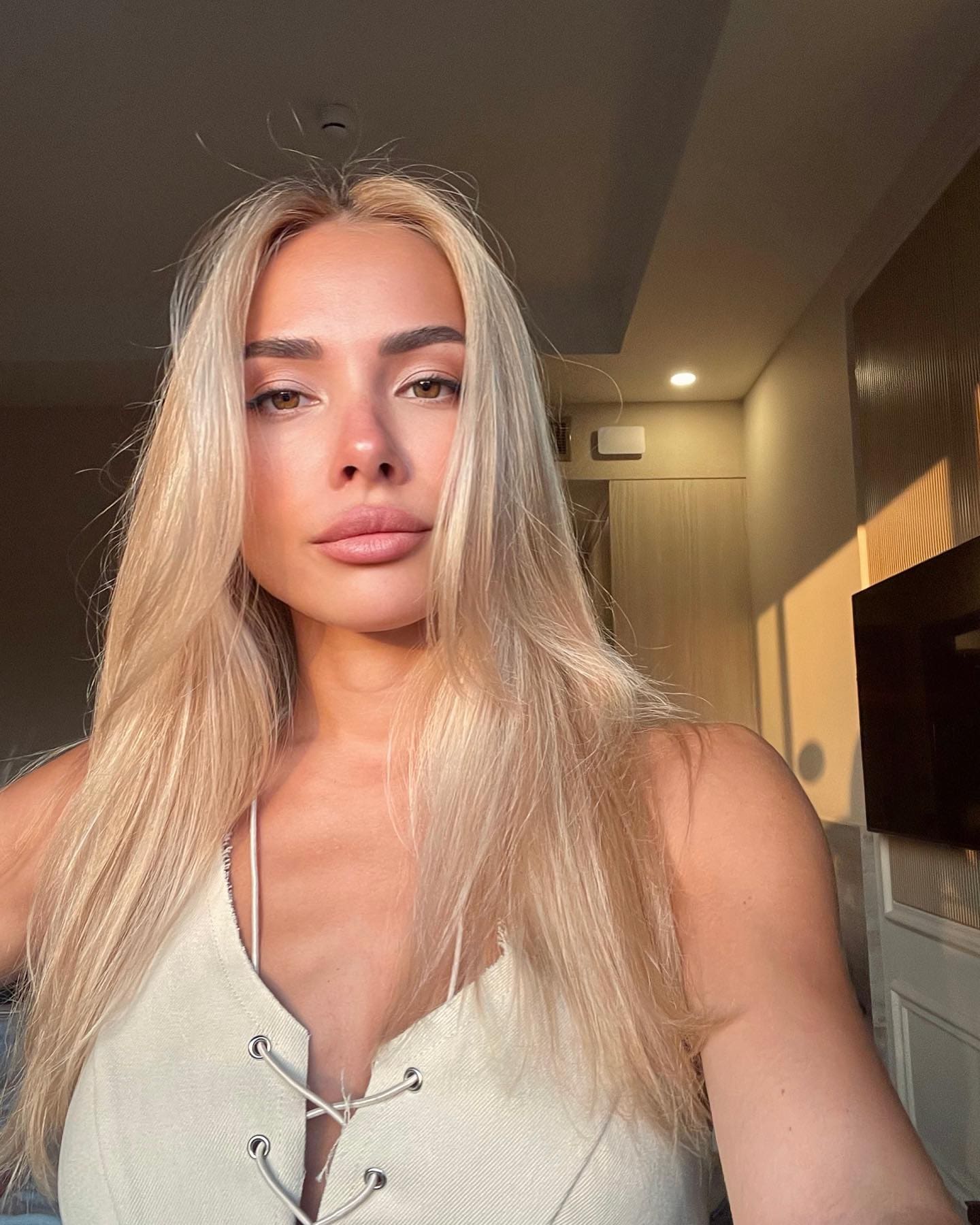 A woman with straight blonde hair takes a selfie in a cream lace-up top, standing indoors near a ceiling light and TV.