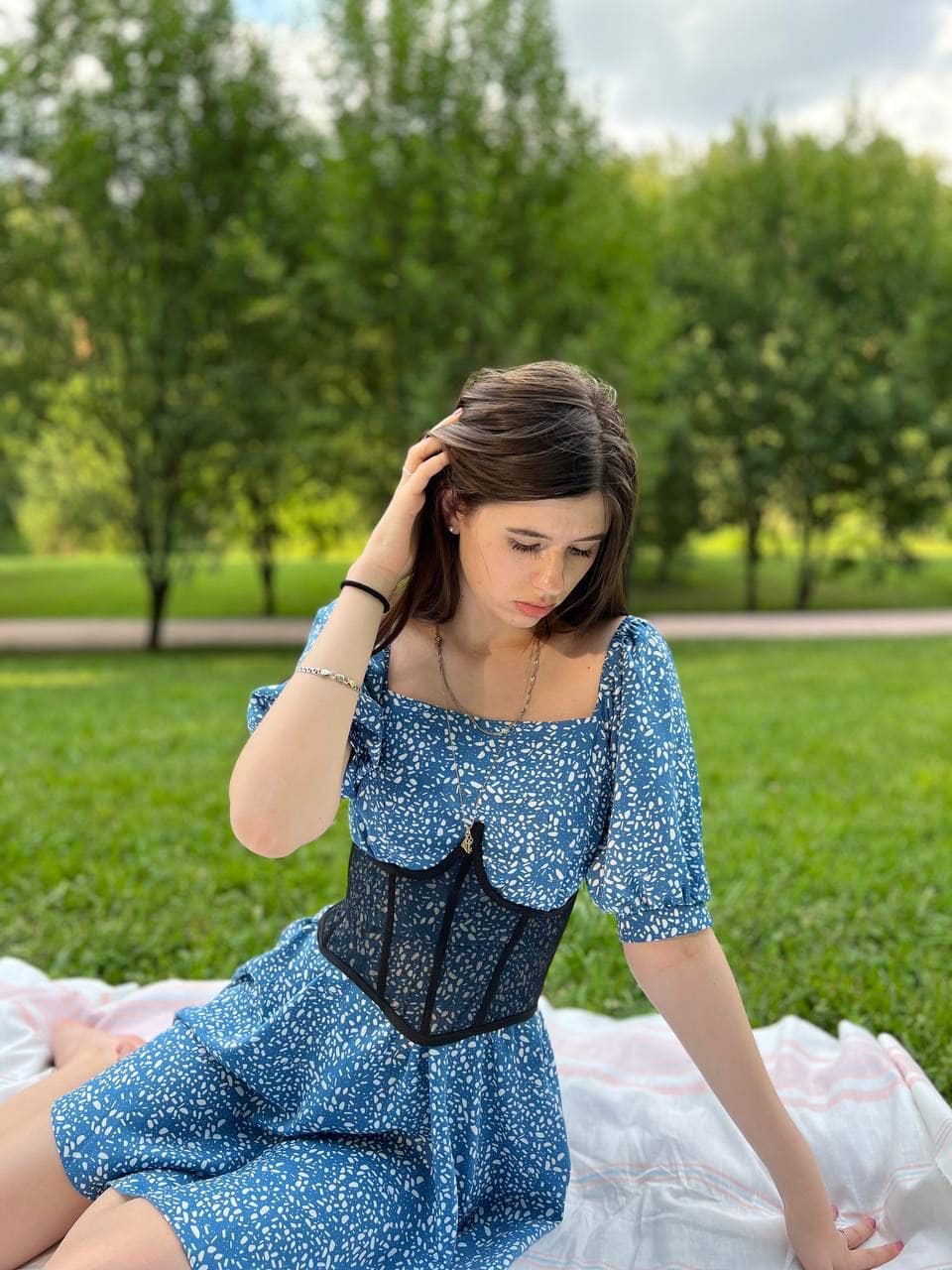 A woman sits on a white blanket in a grassy park, wearing a blue floral dress with a black corset overlay, one hand raised to her hair while looking down.