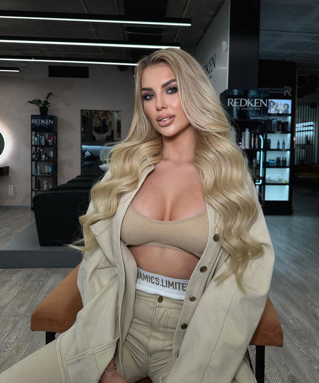 Jeanne sits in a salon chair wearing a beige ribbed crop top, high-waisted trousers, and an open beige jacket, with product shelves behind her.