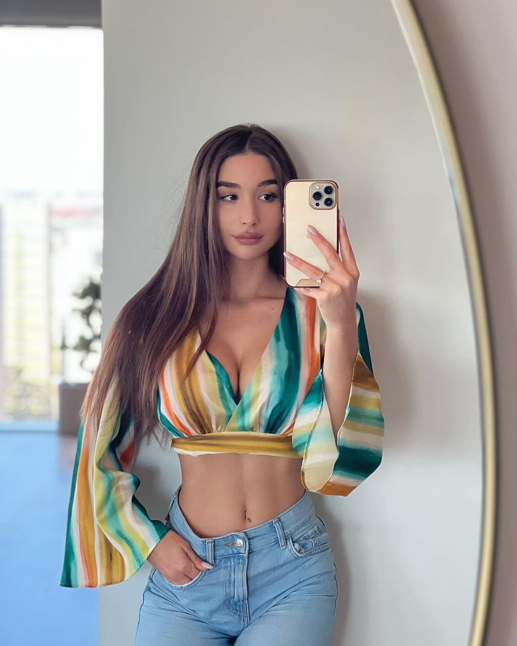 A mirror selfie shows a woman in a striped deep-V crop top and light blue jeans, posing with one hand in her pocket.