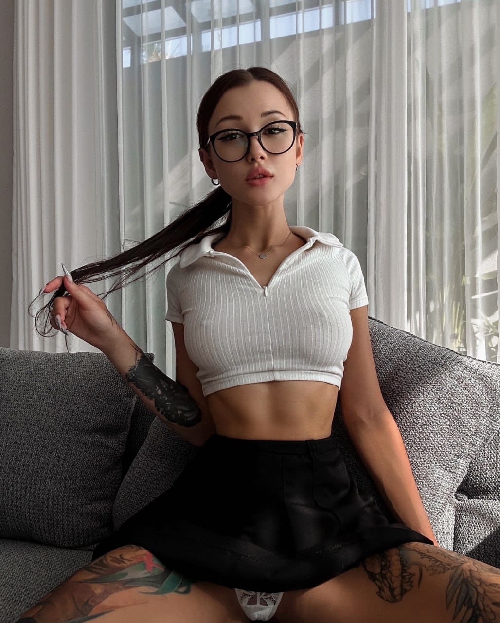 A woman with glasses and tattoos sits on a gray couch in a white crop top and black skirt, holding her hair.