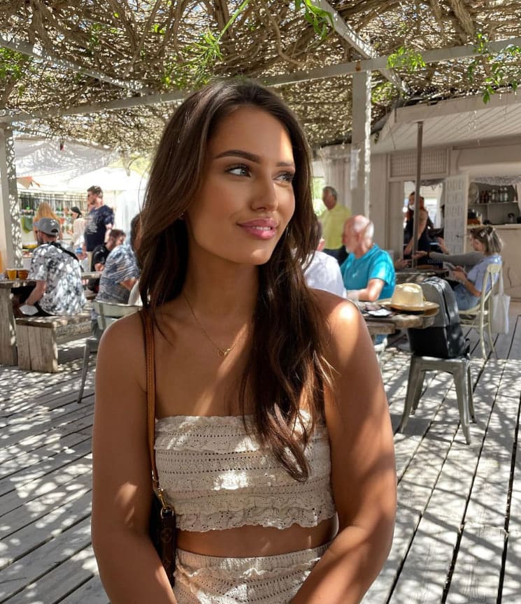 Dana sits on an outdoor wooden deck under a pergola, wearing a cream crocheted crop top and bottoms with a brown shoulder bag.