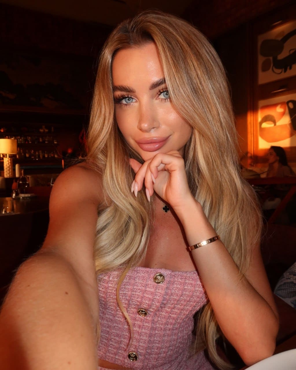 A woman in a pink strapless top rests her chin on her hand while taking a selfie in a dimly lit restaurant.