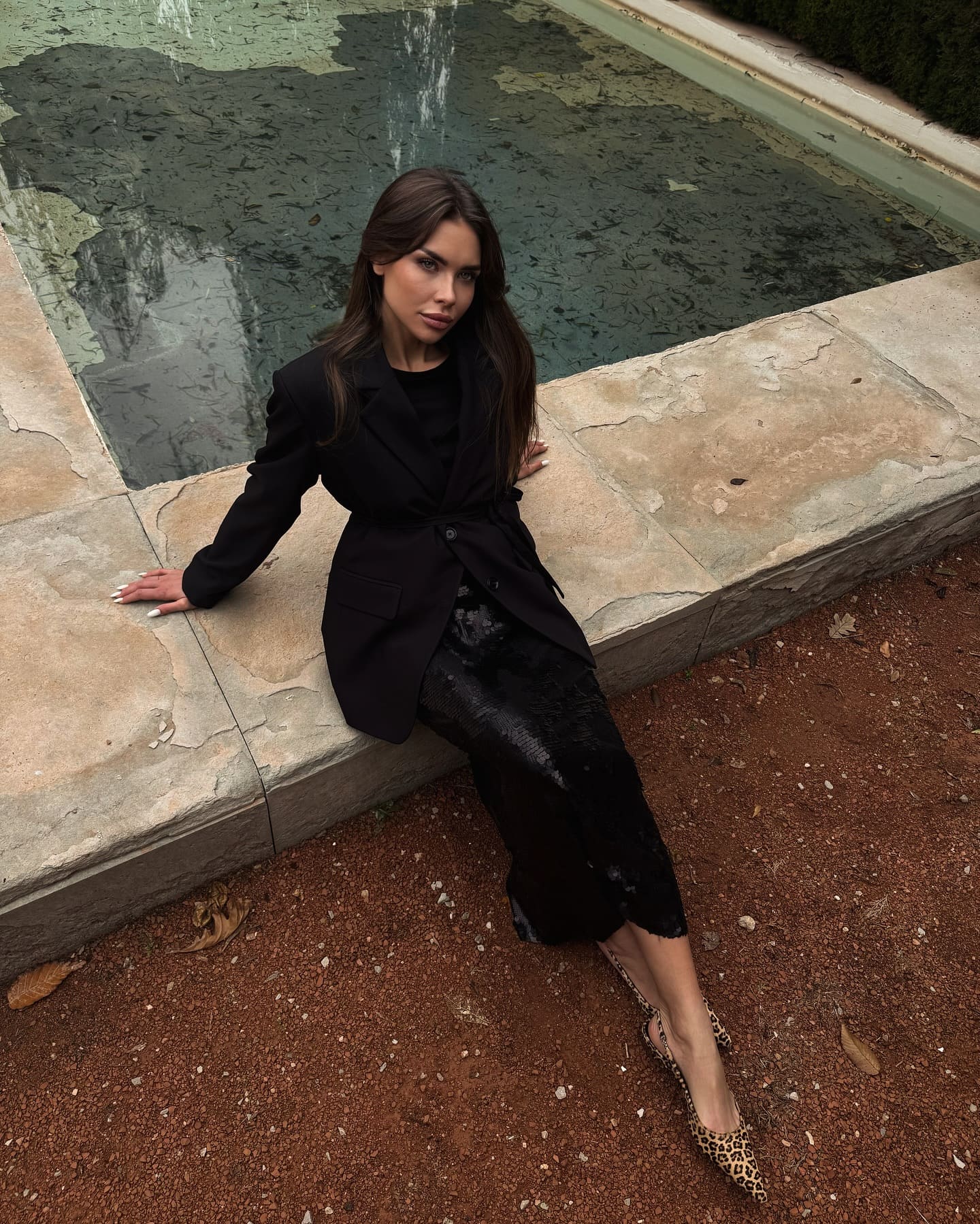 A woman sits on a stone ledge by water wearing a black belted blazer, textured midi skirt, and leopard-print flats.