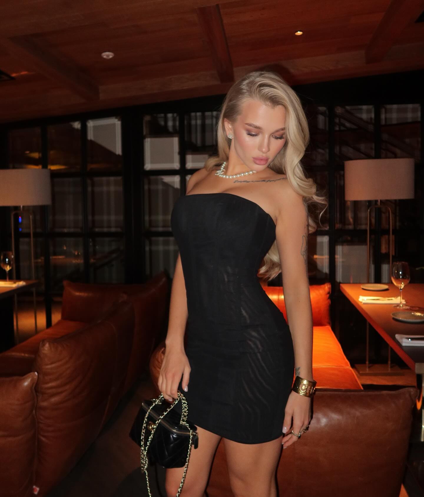 A woman in a strapless black mini dress and pearl necklace stands in a dimly lit restaurant holding a small black handbag.