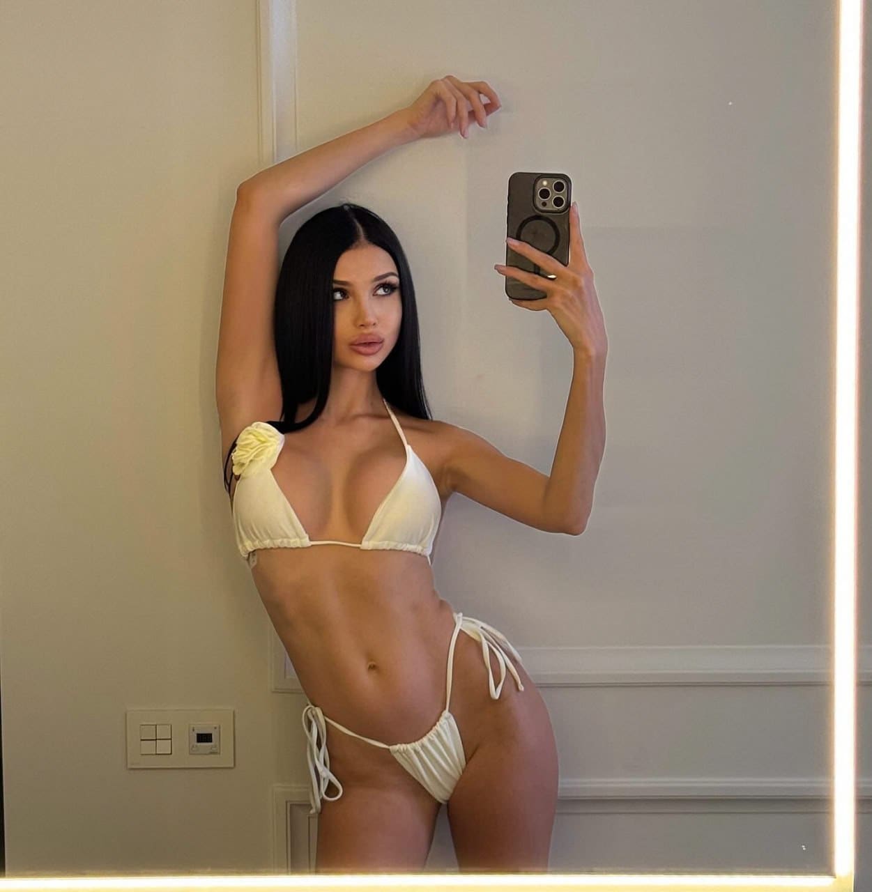 Valeria takes a mirror selfie in a white two-piece bikini with one arm raised, standing against a light wall with a light switch and vertical light strip.