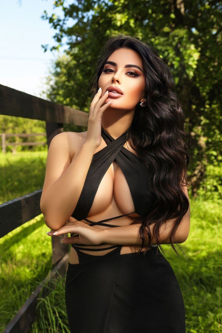 A woman in a black strappy halter top and high-waisted skirt stands by a wooden fence, hand raised to her chin.