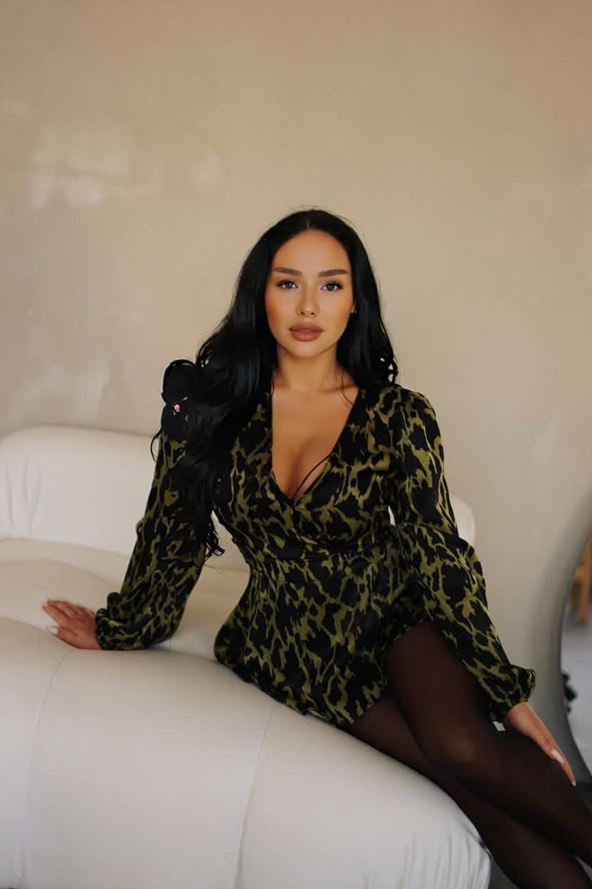 Olya sits on a white curved sofa wearing a green and black animal print mini dress with a deep V-neck and black sheer tights.